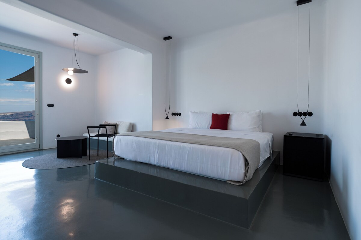 Un dormitorio moderno con una cama grande, mesitas de noche, una silla y vistas a través de un ventanal amplio.