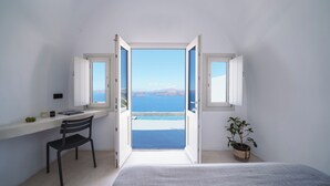 Une chambre à coucher avec un lit, un bureau et une chaise, une plante en pot, et une vue sur la mer par une porte ouverte.