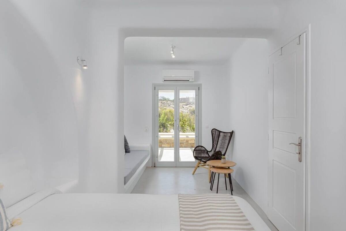 A minimalist bedroom with a bed, a chair, a small table, and a door leading to a balcony.