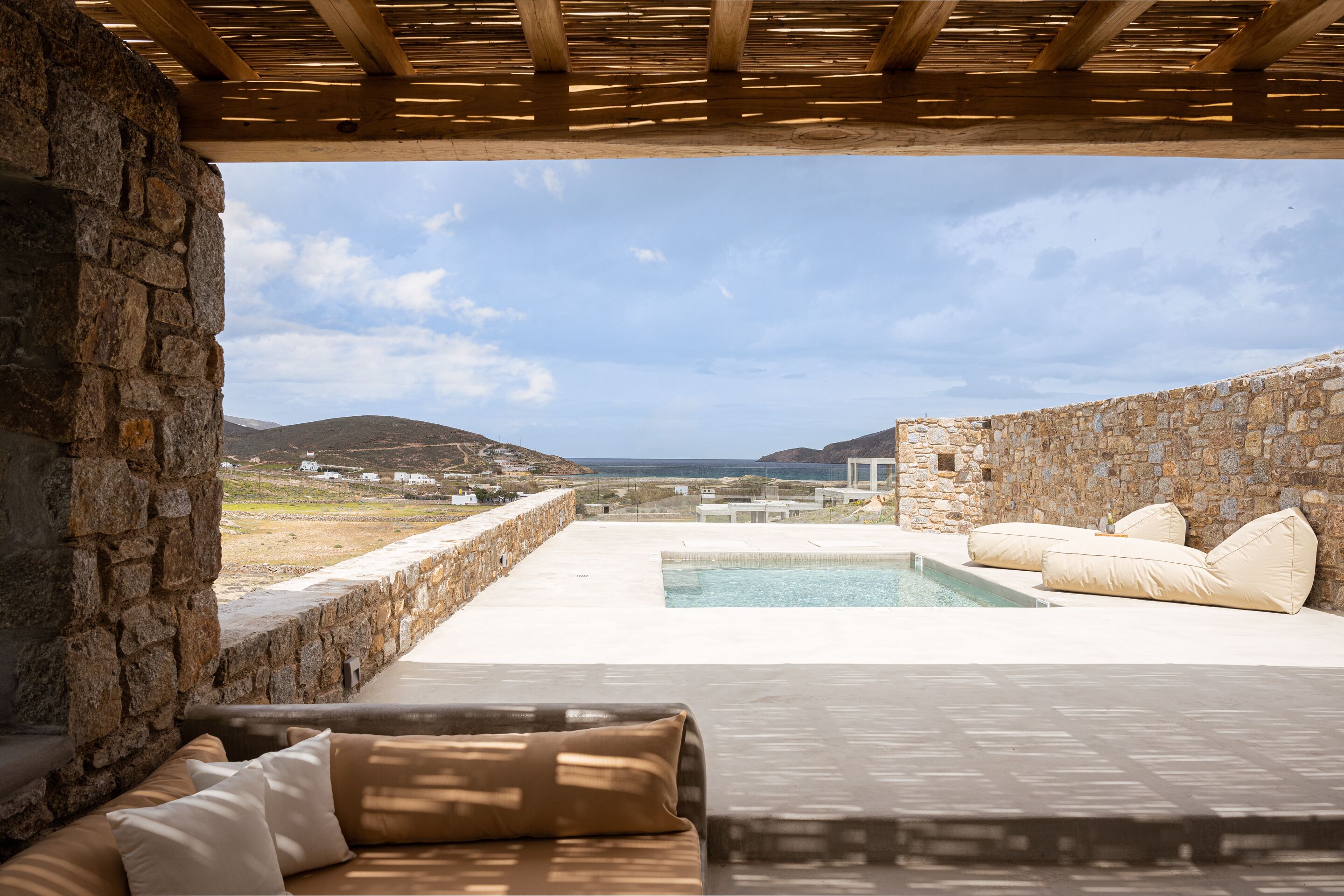 A stone-walled outdoor area with a pool, a wooden pergola, and a sofa with cushions.