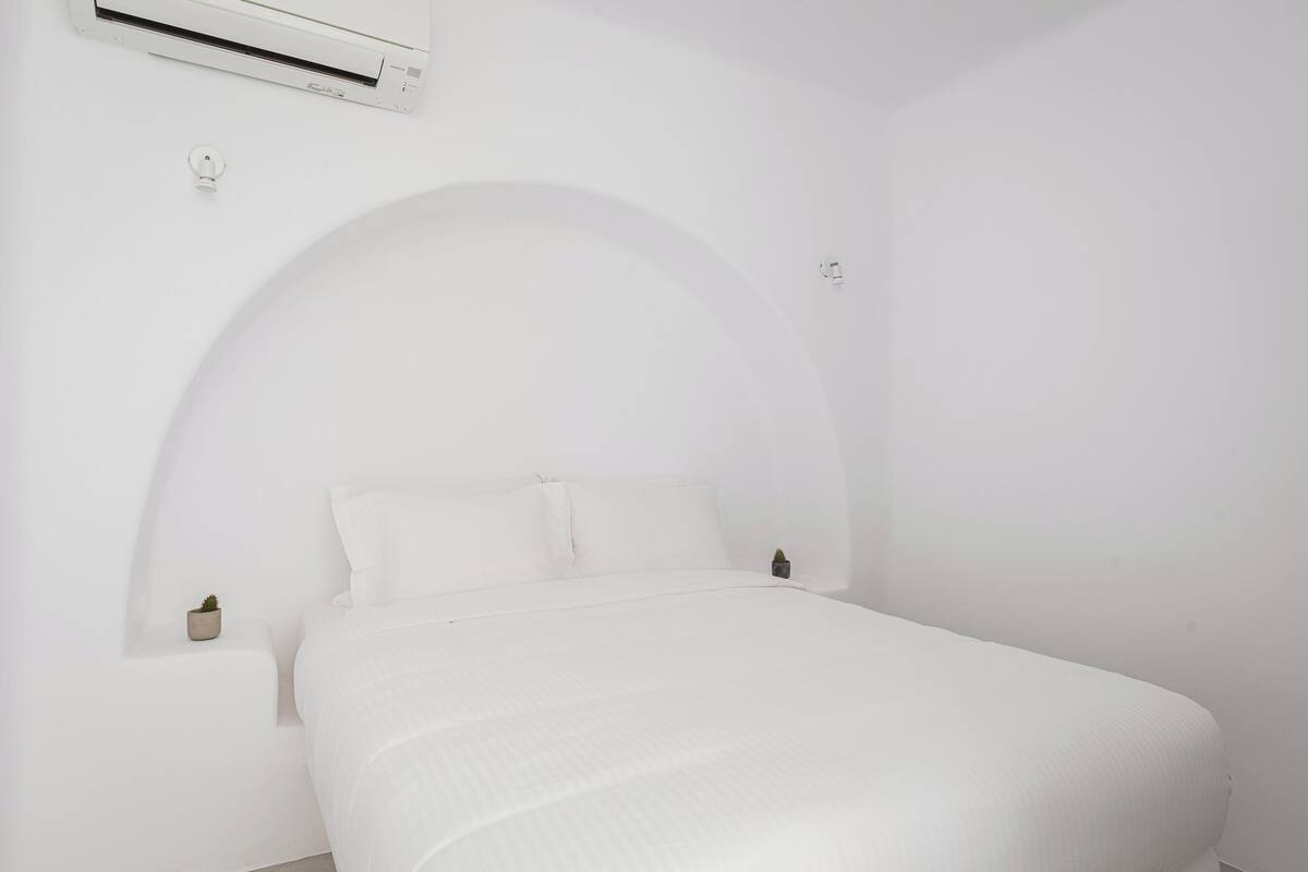 A white room with a bed, an air conditioner, and a small plant on the nightstand.