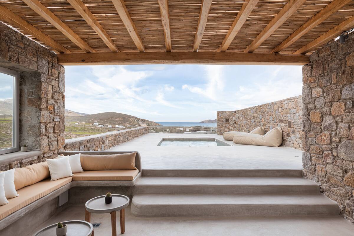 A stone-walled outdoor seating area with a pool, wooden ceiling, and a view of the sea.