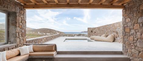 A stone-walled outdoor seating area with a pool, wooden ceiling, and a view of the sea.
