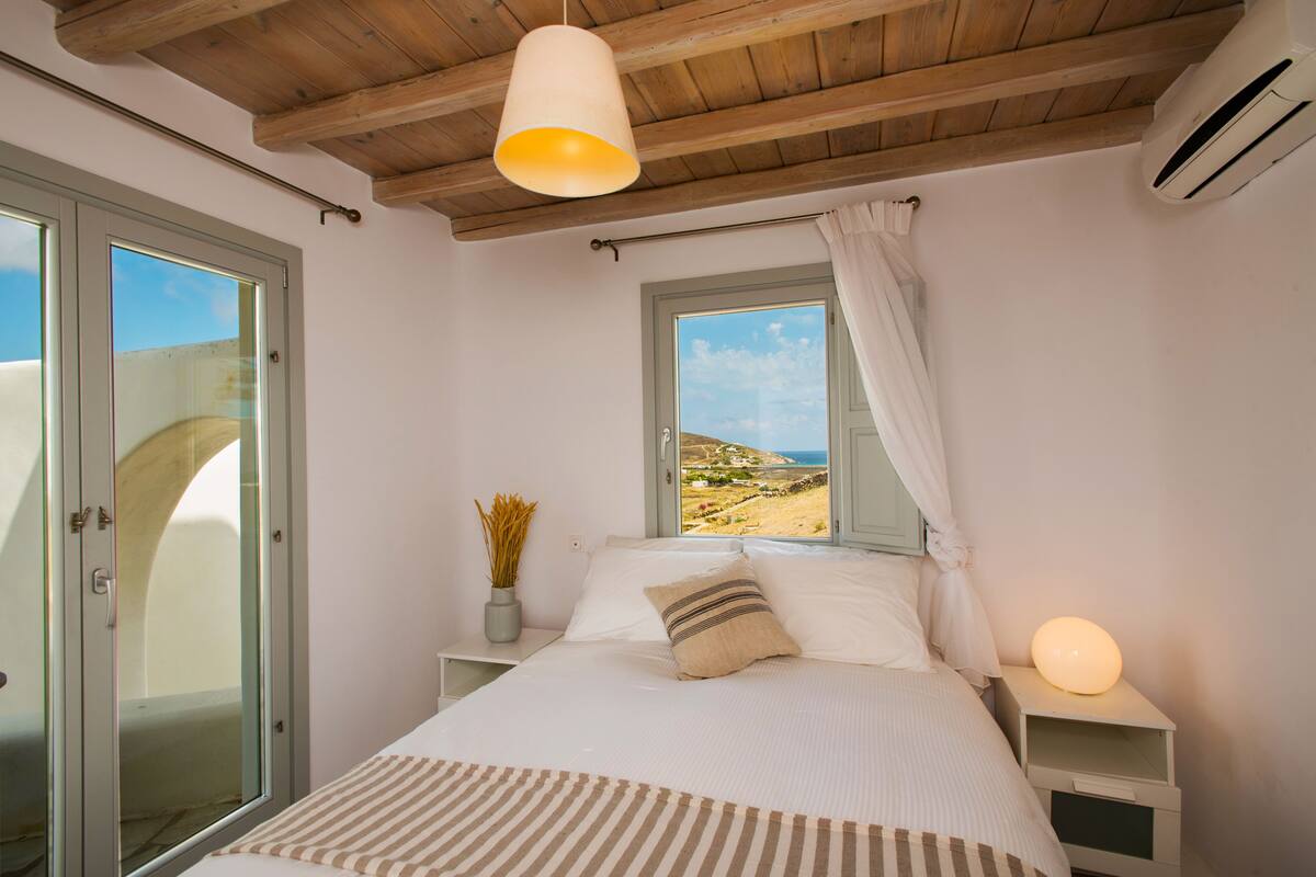 A bedroom with a bed, a bedside table, a nightstand, a window with a view, and a ceiling fan.