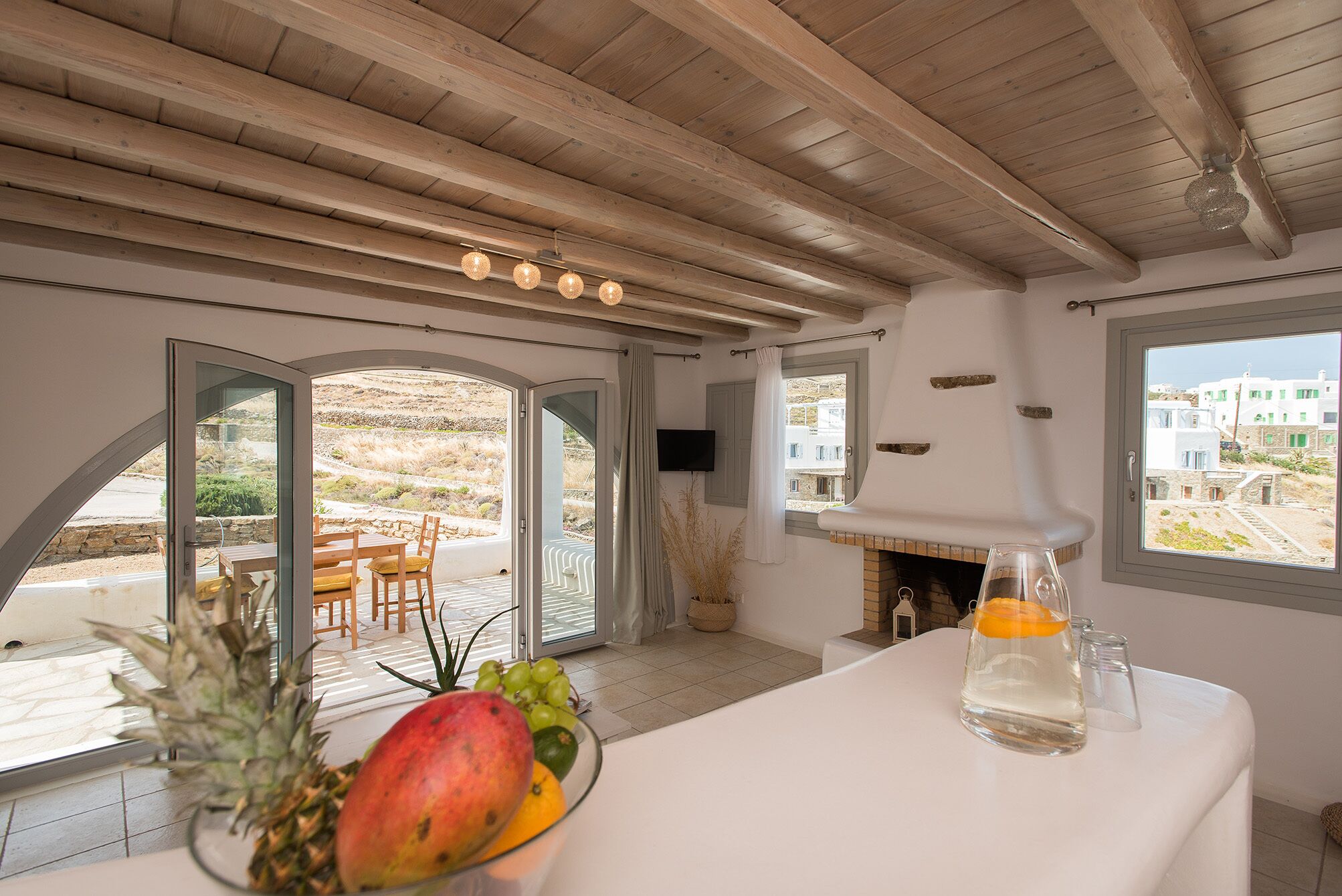 Une pièce avec un plafond en bois, une cheminée, une table avec des fruits et un verre d’eau, et une vue sur les bâtiments à travers de grandes fenêtres.