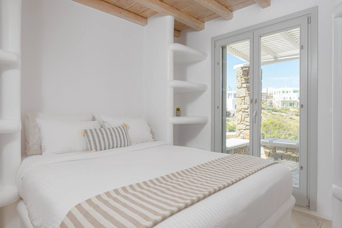 A bedroom with a bed, white walls, a stone pillar, and a view of a balcony with outdoor furniture.