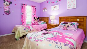 Habitación con dos camas, cada una con una colcha con motivos de Minnie Mouse, un cabecero de madera, una mesita de noche con lámpara y una ventana con cortinas rosadas.