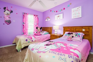 A room with two beds, each with a Minnie Mouse themed bedspread, a wooden headboard, a nightstand with a lamp, and a window with pink curtains.