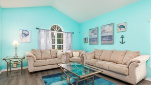 A living room with two beige sofas, a glass coffee table, a blue patterned rug, and nautical-themed wall art. - Luxury 5 bed 4 bath SW facing pool with conservation and lake view (Clermont)