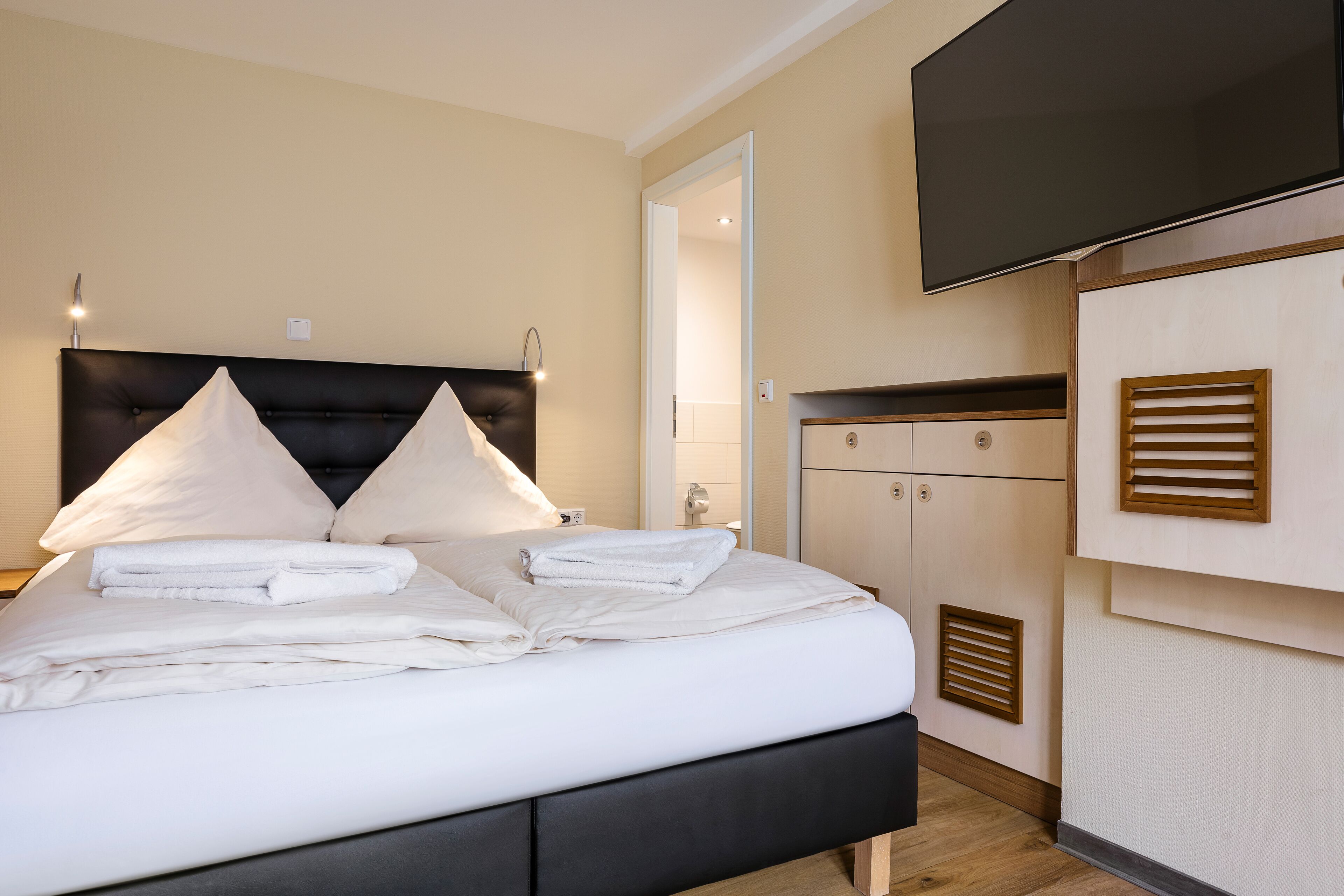 Een opgemaakt bed met witte lakens en kussens, een zwart hoofdbord en een flatscreen televisie erboven.