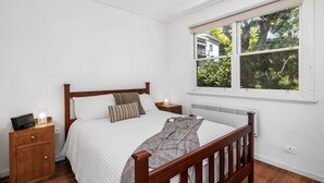 A bedroom with a wooden bed, white walls, a window with a view of trees, and a bedside table with a lamp.