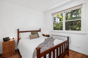 A bedroom with a wooden bed, white walls, a window with a view of trees, and a bedside table with a lamp.