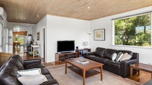 Uma sala de estar moderna com teto de madeira, uma janela ampla, uma TV de tela plana, um sofá de couro, uma mesa de centro e uma estante de livros.