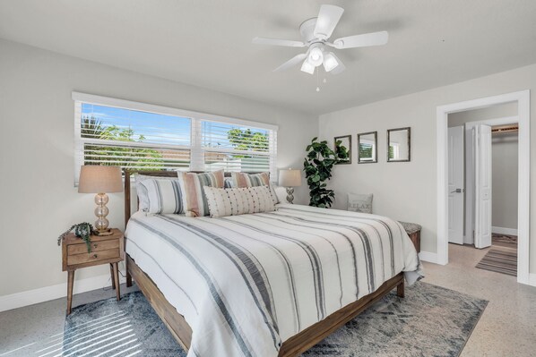 A bedroom with a bed, nightstands, a ceiling fan, a window with blinds, and a door leading to another room.