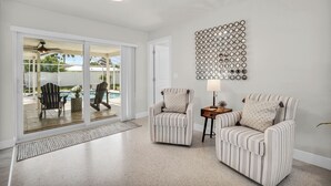 Un salón espacioso con un gran ventanal corredero que da a una zona de piscina, dos sillones a rayas, una mesita con una lámpara y un adorno en la pared.