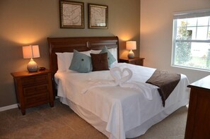 A neatly made bed with white linens, two pillows, and a brown throw blanket. There are two bedside tables with lamps, a window with a view of greenery, and two framed pictures on the wall.