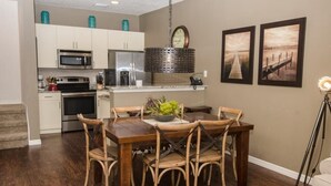 A dining area with a wooden table and chairs, a kitchen with white cabinets and stainless steel appliances, and two framed pictures on the wall.