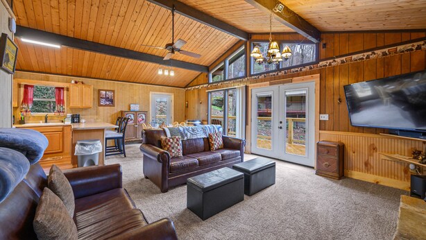 A spacious living area with a leather sofa, a coffee table, and a television. There is a kitchen area with a window and a ceiling fan.