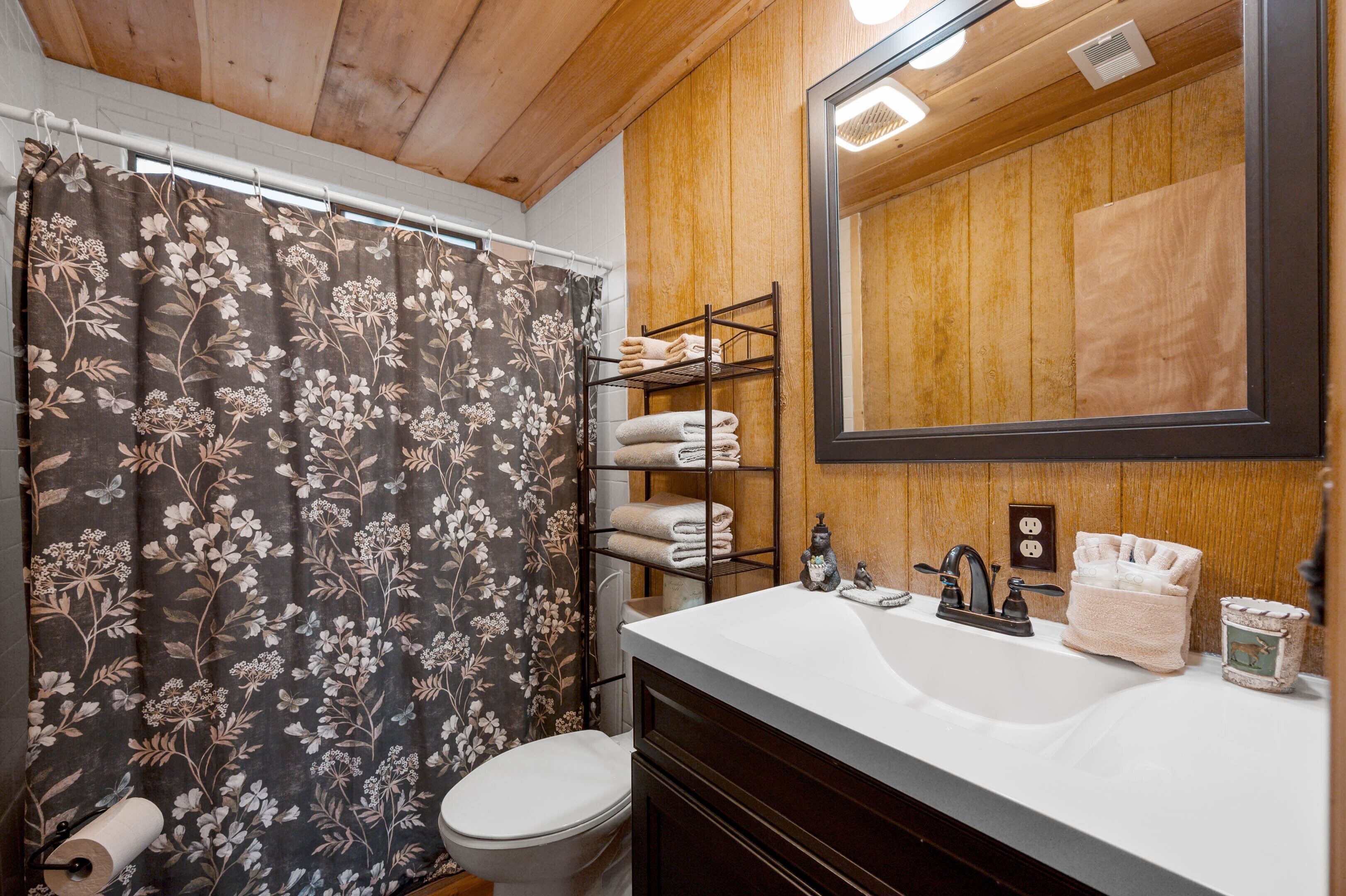 Un baño con techo de madera, cortina de ducha con estampado floral, lavamanos blanco con espejo arriba y repisa con toallas.