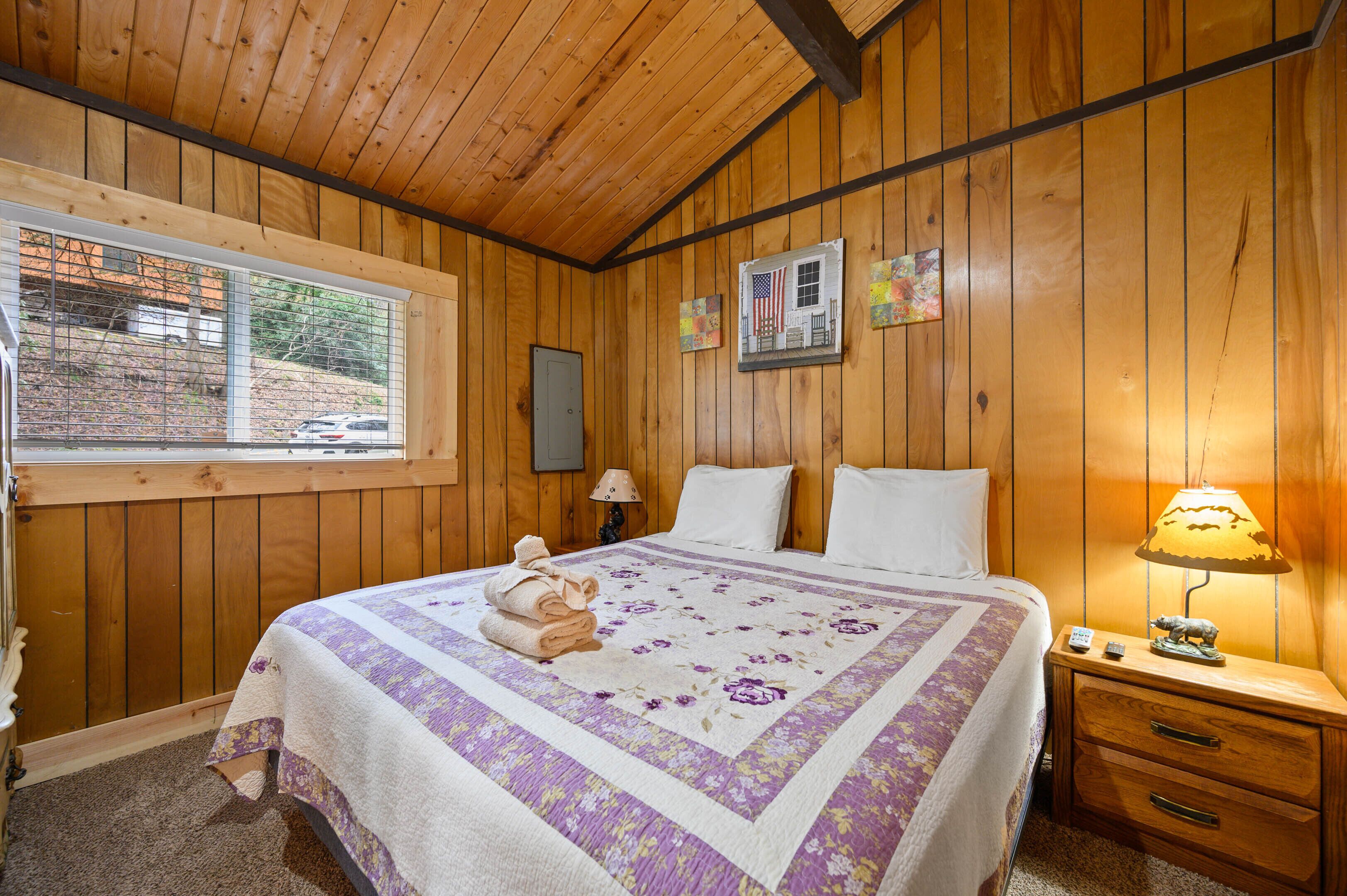 A wooden-paneled bedroom with a bed, a nightstand, a window, and a wall-mounted lamp.