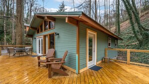 A wooden deck with a dining area and chairs, a small building with a door and windows, surrounded by trees.
