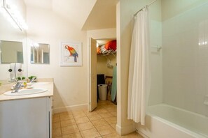 Een badkamer met een douche, een wastafel en een spiegel. Een deur leidt naar een inloopkast met kleding en een prullenbak.