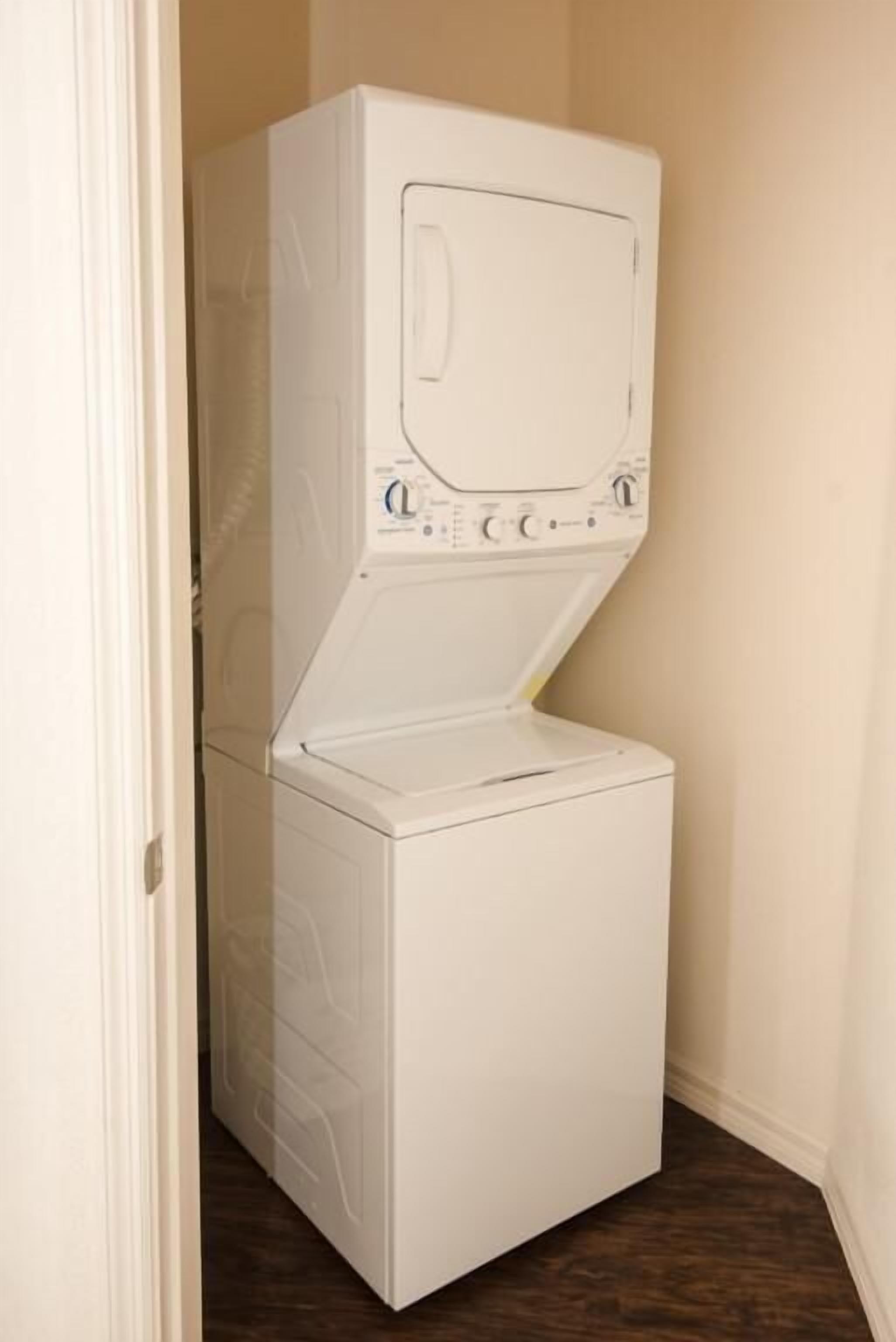 A white stacked washer and dryer unit in a corner of a room with wooden flooring.