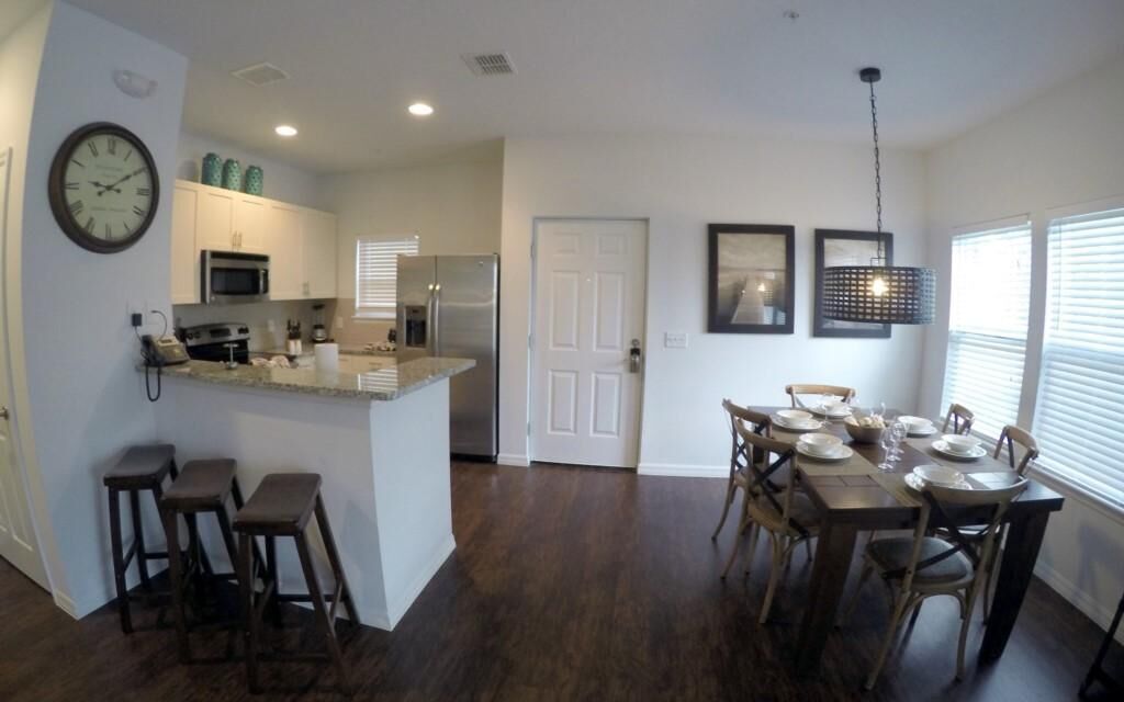 A modern kitchen with a central island, bar stools, a dining table set for six, and a large clock on the wall.