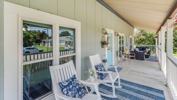 Eine überdachte Veranda mit weißen Schaukelstühlen und einem Tisch, einer blau-weißen Streifenoptik als Teppich und Blick auf ein Wohngebiet.