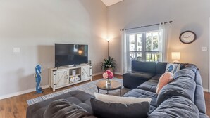 Ein Wohnzimmer mit einer großen Couch, einem Fernseher, einem kleinen Tisch mit Blumen und einem Fenster mit Vorhängen.