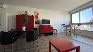 A modern living room with a dining area, a TV, and a large window. - Sol-y-Days Murènes 1, large studio close to beaches and center (Le Grau-du-Roi)