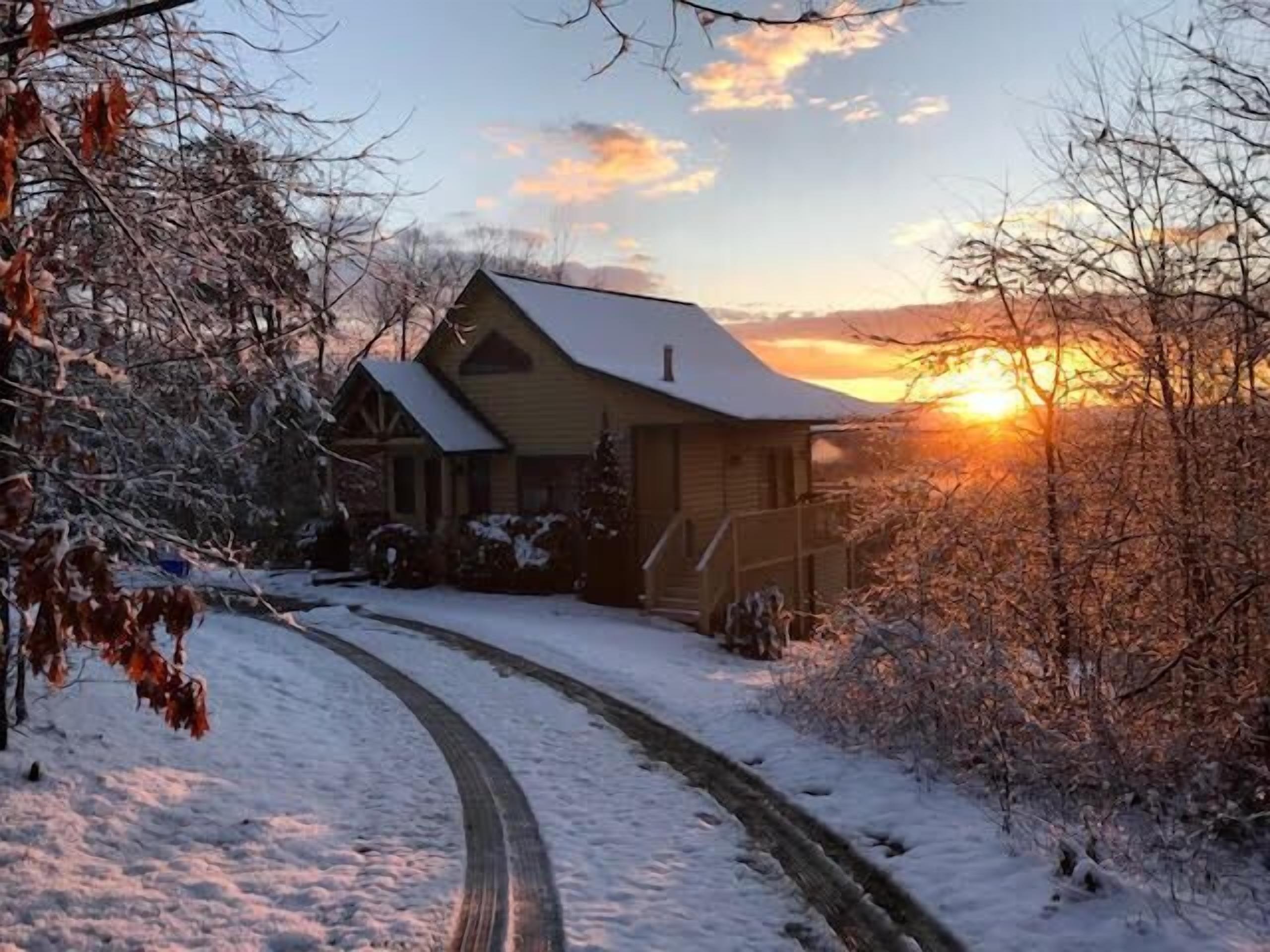 A snow-covered house with a wooden exterior and a sloped roof, surrounded by trees and a clear sky.