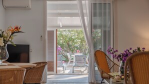 Una habitación con una mesa de comedor, sillas, un televisor y un ventanal corredero que da a un área exterior con sillas y plantas.
