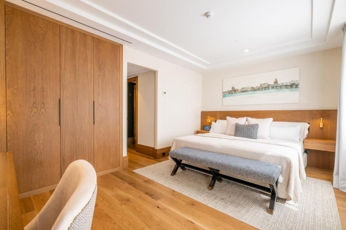 A bedroom with a large bed, a wooden wardrobe, a bench, and a cityscape painting on the wall.