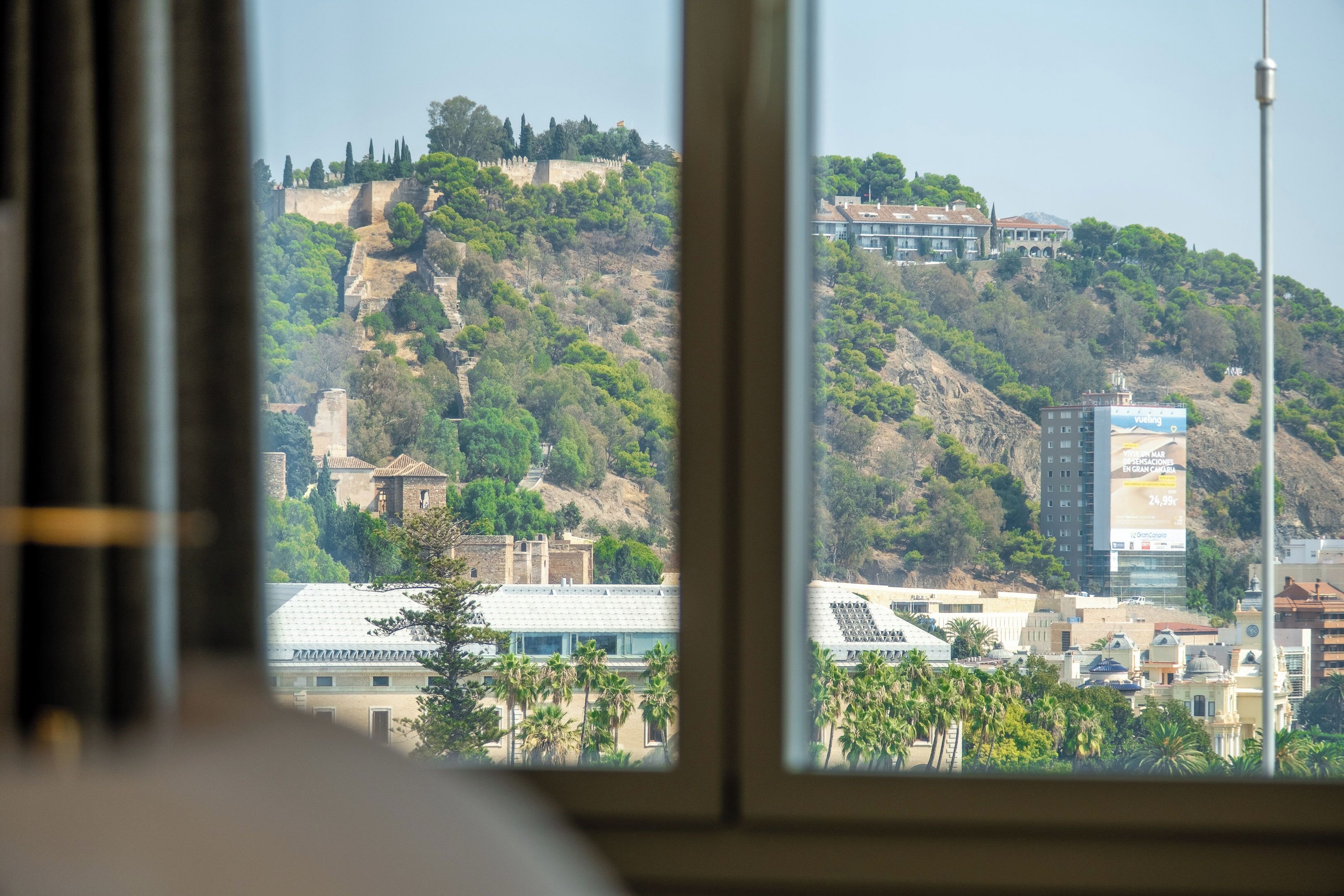 Vista a través de una ventana mostrando un paisaje urbano con una colina, edificios y un anuncio.