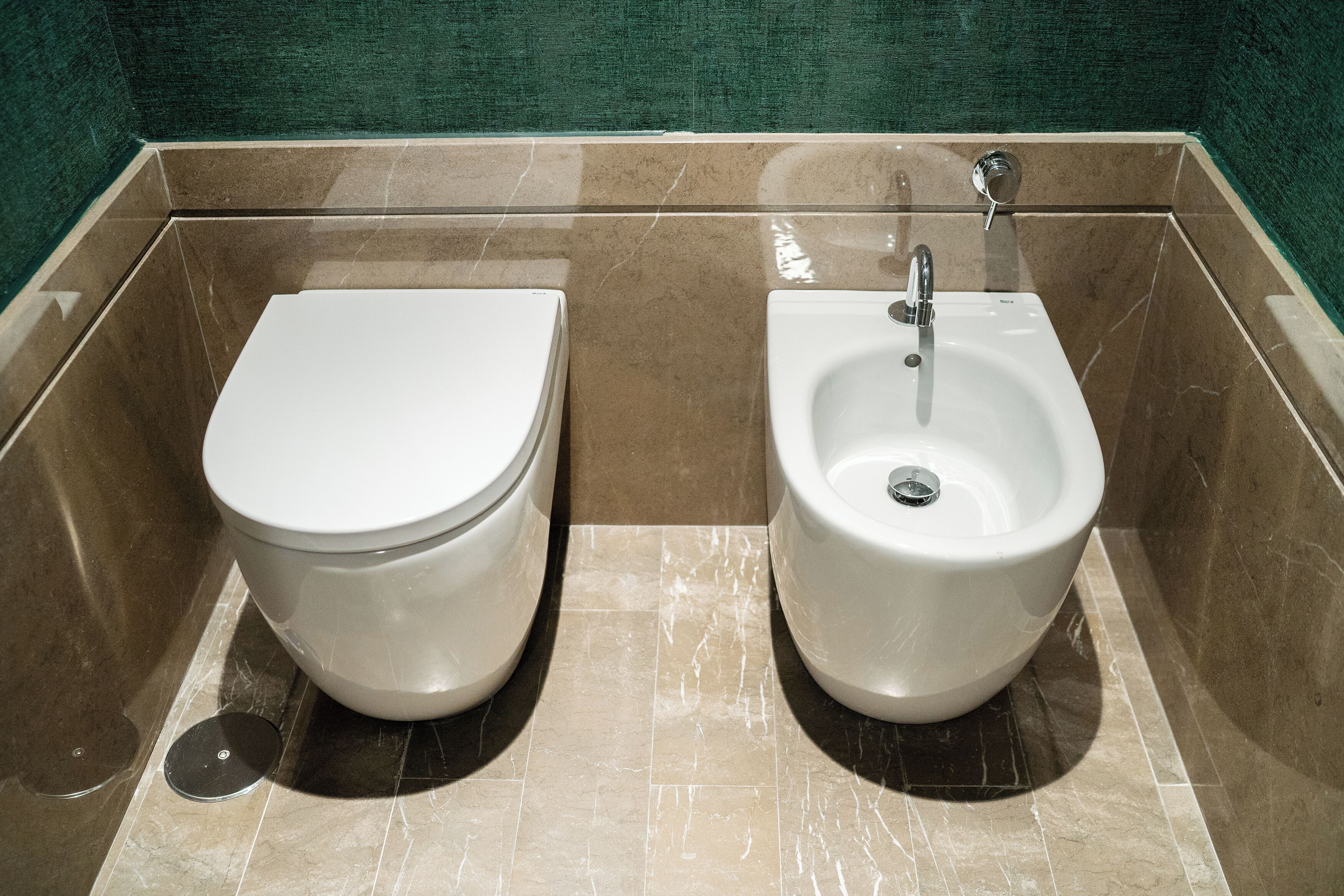 Ein Badezimmer mit Toilette und Bidet, beide weiß, in einem mit Fliesen ausgelegten Bereich. Im Hintergrund ist eine grüne Wand zu sehen.