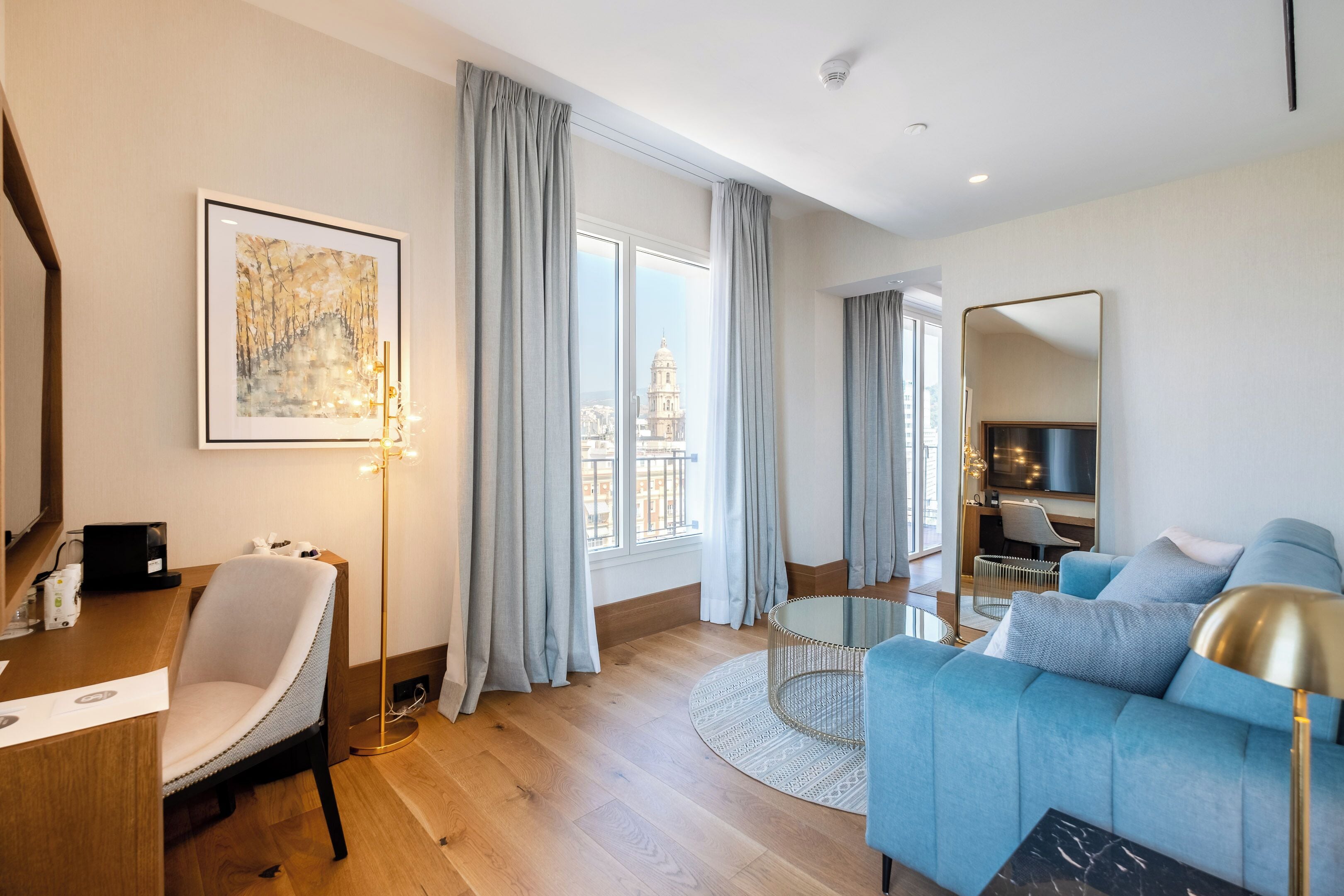 Una sala de estar moderna con un sofá azul, una mesa de centro redonda y un escritorio con una silla. Se ve un ventanal con cortinas, una obra de arte enmarcada en la pared y un espejo.