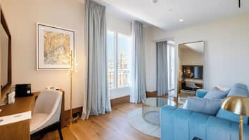 Una sala de estar moderna con un sofá azul, una mesa de centro redonda y un escritorio con una silla. Se ve un ventanal con cortinas, una obra de arte enmarcada en la pared y un espejo.