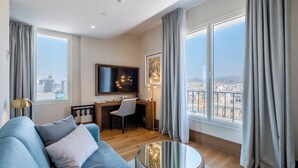 A modern living room with a sofa, a desk, and a TV. There are large windows with curtains, and a city view is visible. - Soho Boutique Equitativa (Málaga)