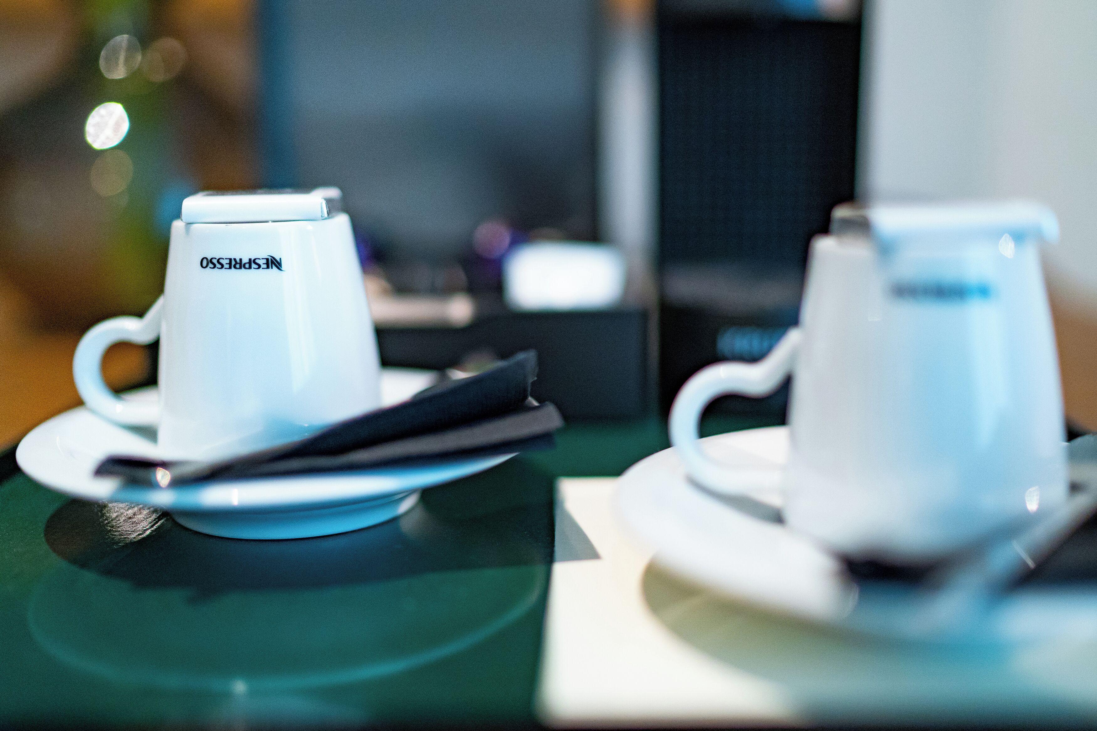 Two white coffee cups with lids on saucers, placed on a table with a blurred background.