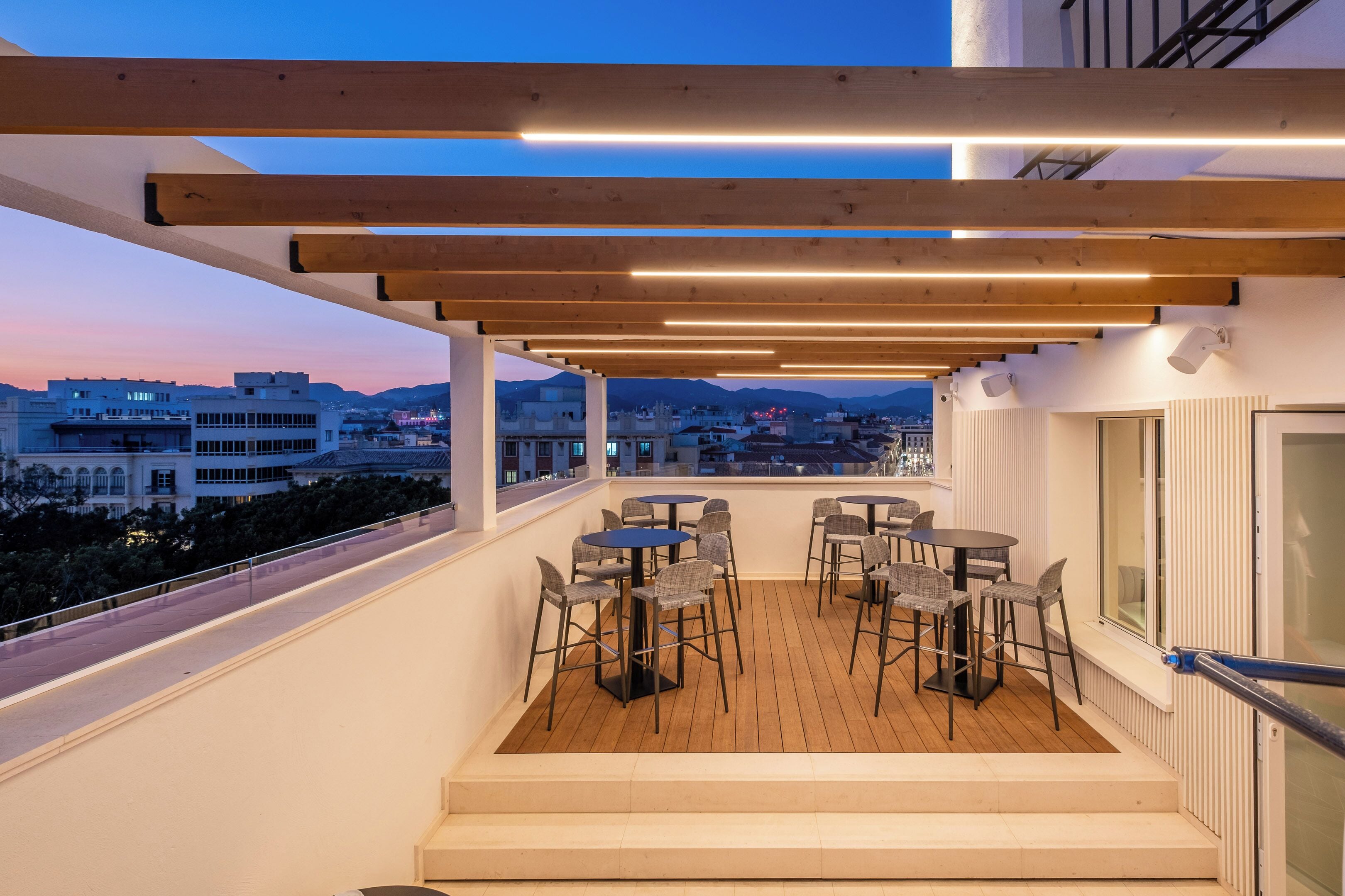 Un balcon doté d’un plancher en bois, de sièges pour l’extérieur et offrant une vue sur la ville au crépuscule.