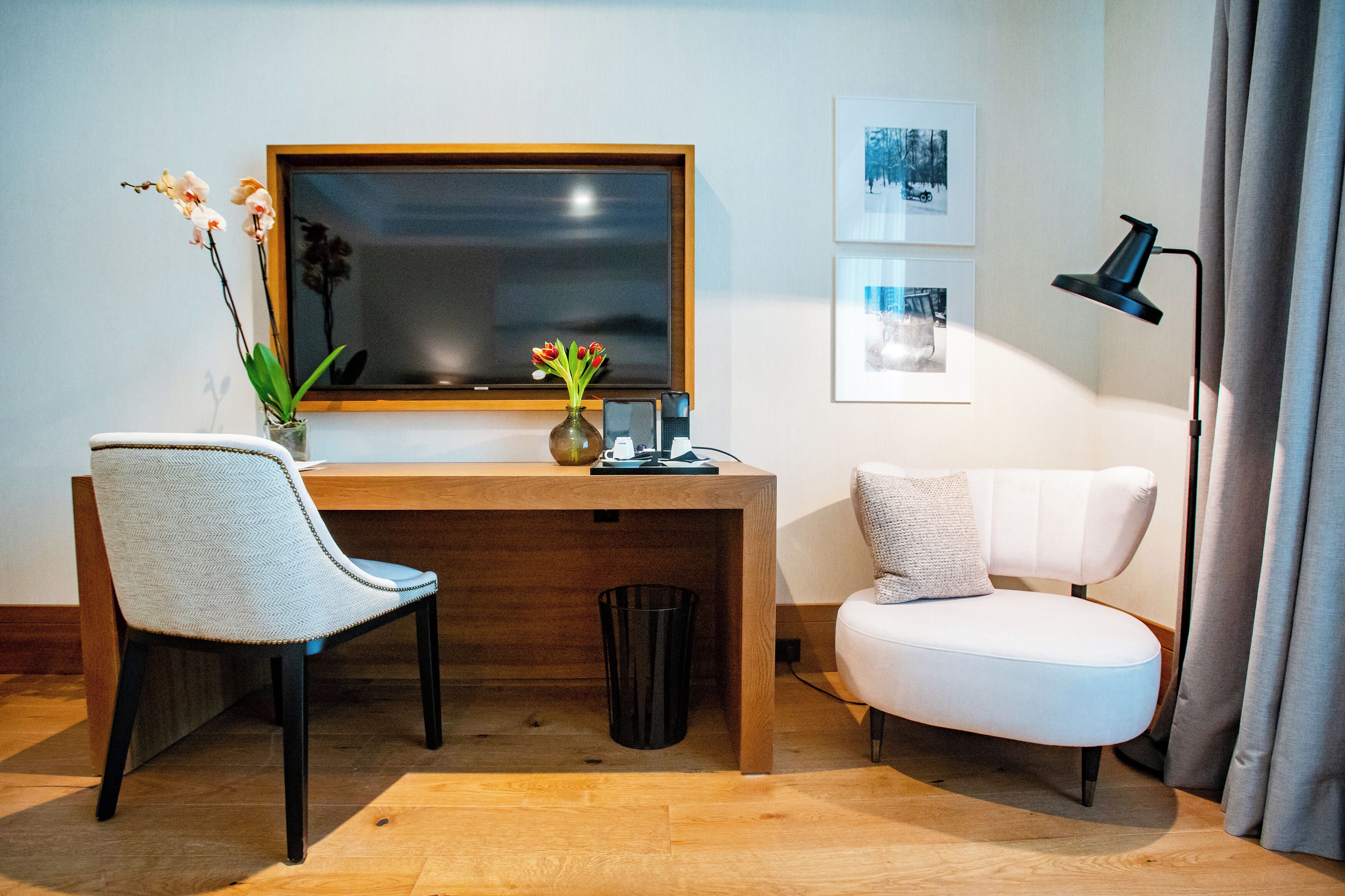 Una habitación moderna con un escritorio de madera, una silla blanca, un pequeño jarrón con flores, un cesto de basura negro y una lámpara de pie negra.