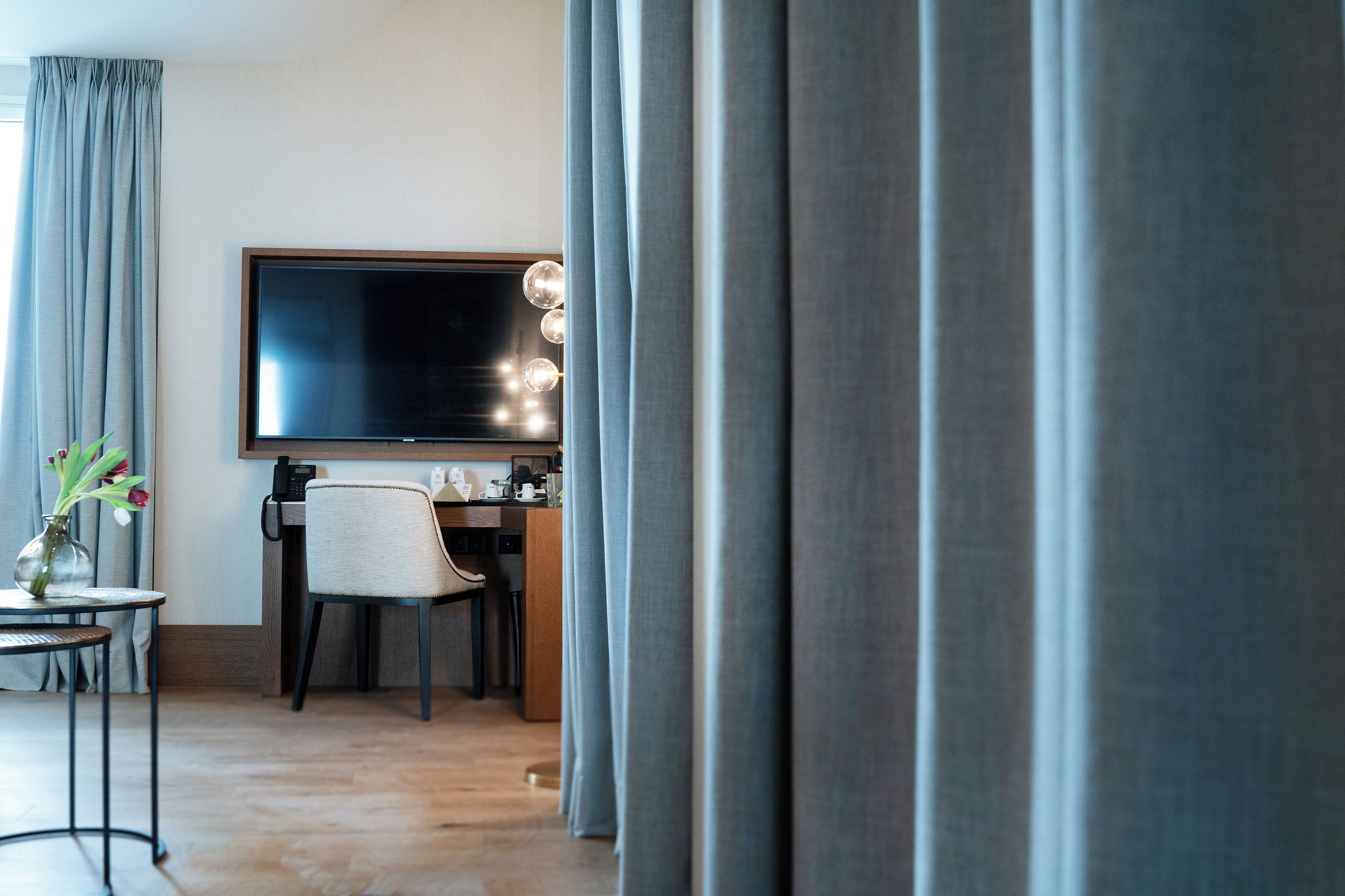 Ein modernes Hotelzimmer mit einem großen Flachbildfernseher, einem Schreibtisch mit Stuhl und einem kleinen Tisch mit Blumenvase.