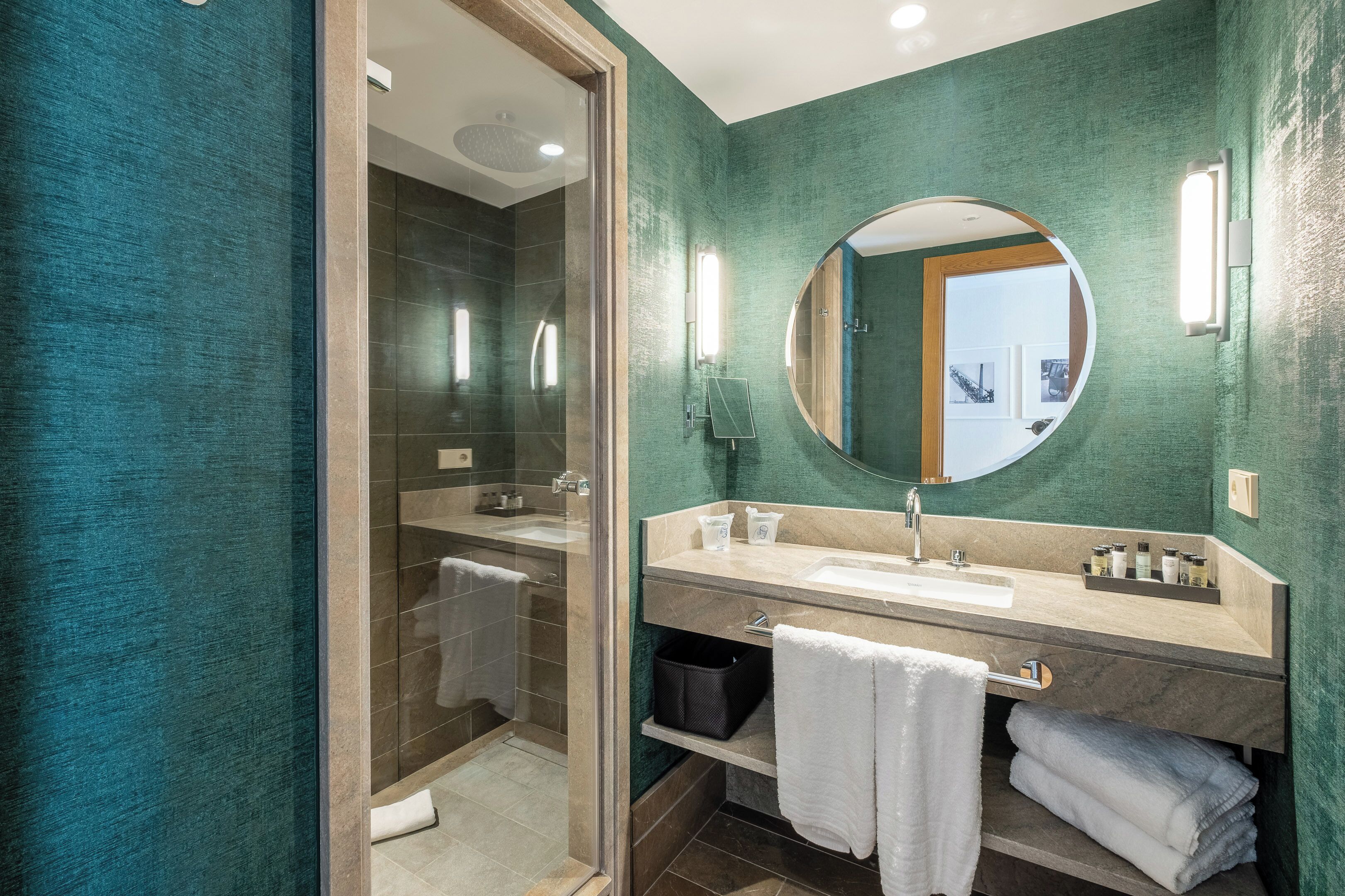 Un baño moderno con ducha de vidrio, un espejo redondo, un lavamanos con tocador y una pared verde con textura.