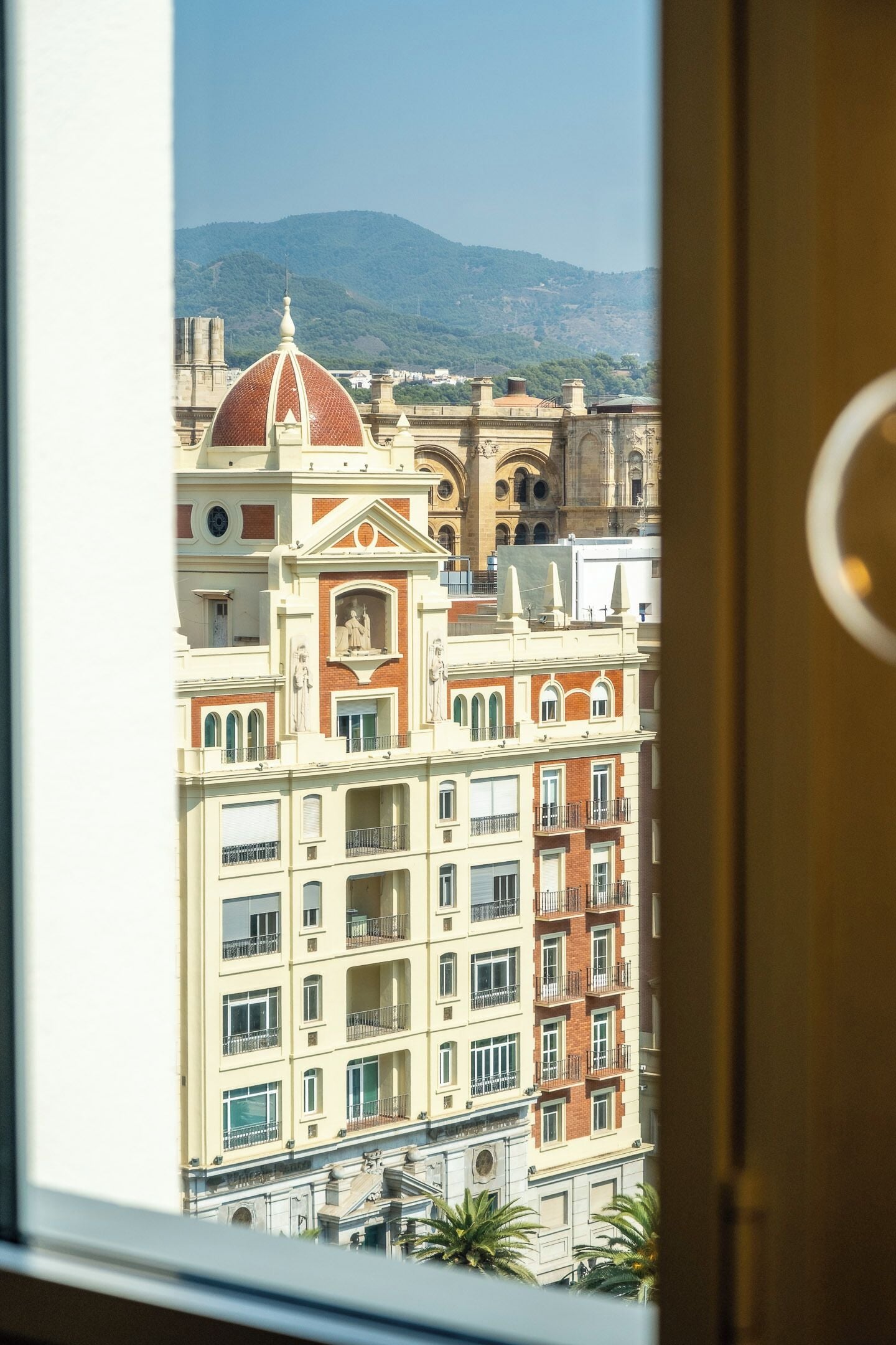 Vista de um edifício com vários andares, uma cúpula imponente e arquitetura clássica, vista através de uma janela com design circular.