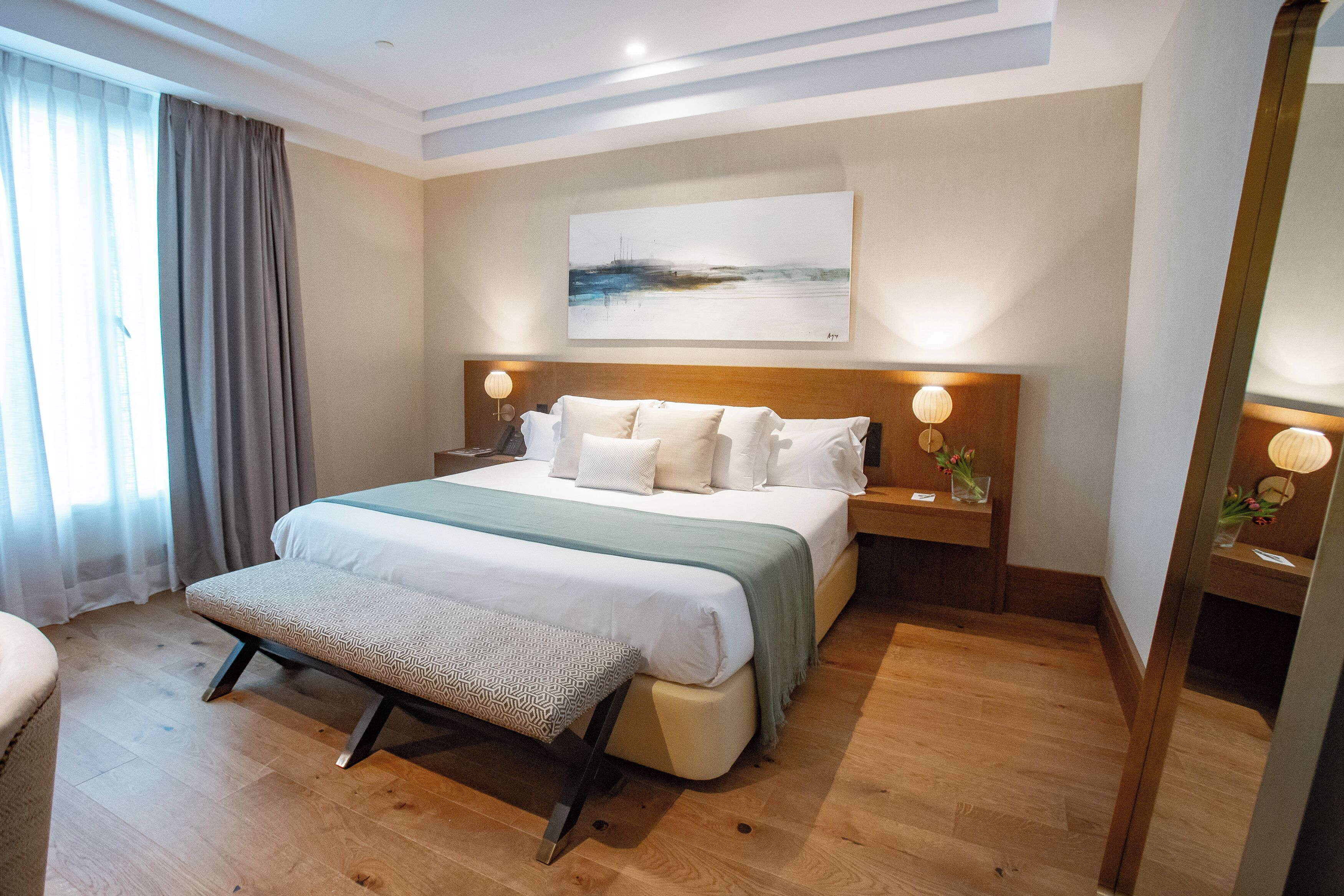 A modern hotel room with a large bed, a bedside table with a lamp, a bench, and a large window with curtains.