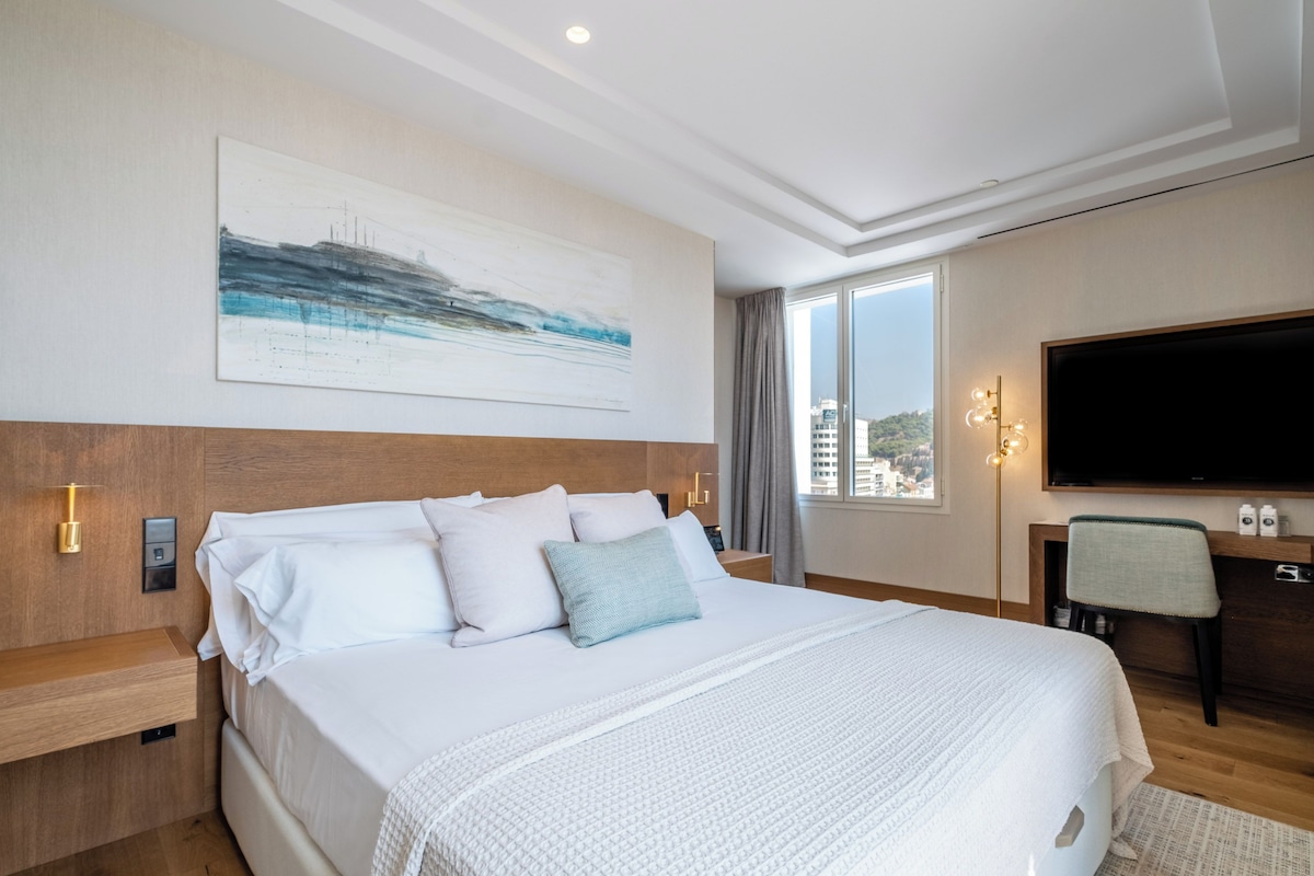 A modern hotel room with a large bed, a flat-screen TV, a desk, and a city view.