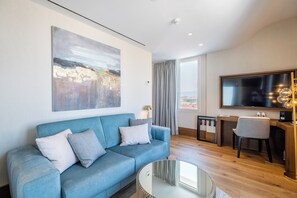 A modern living room with a blue sofa, a glass coffee table, a large abstract painting, a flat-screen TV, and a view of the city through large windows. - Soho Boutique Equitativa (Málaga)