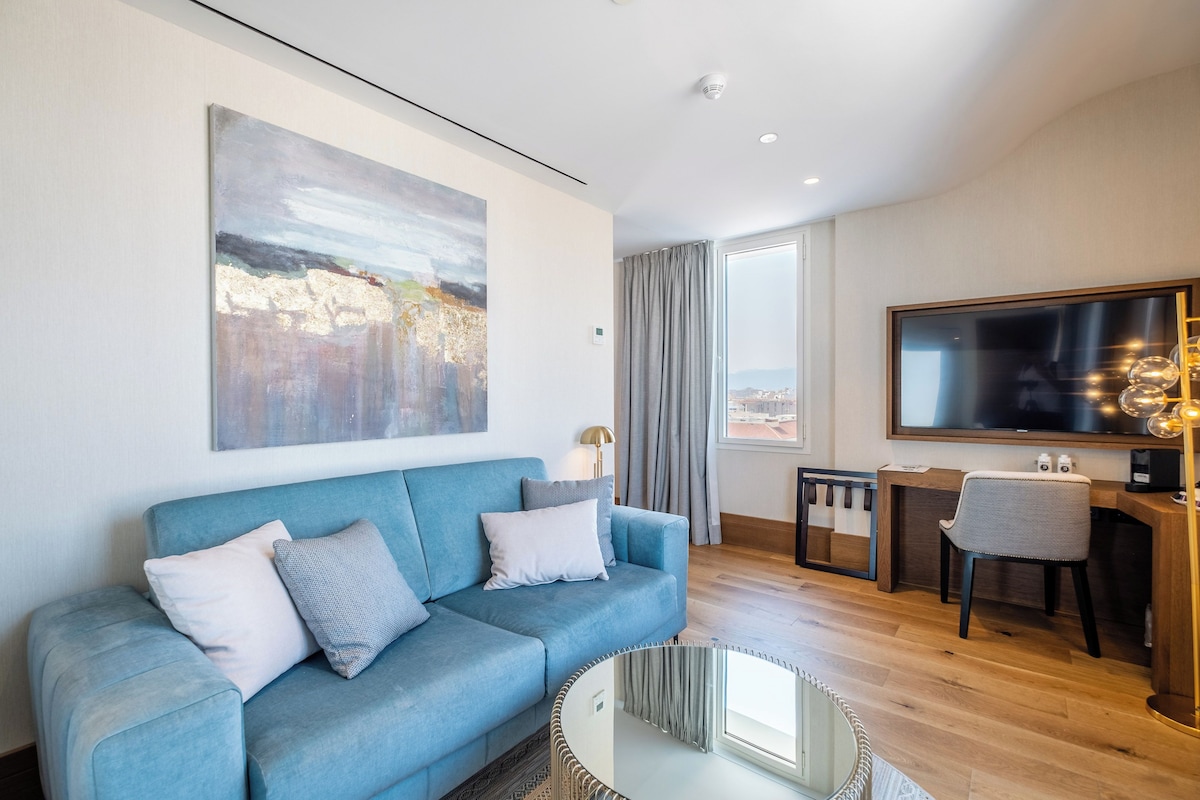 A modern living room with a blue sofa, a large abstract painting, a flat-screen TV, and a dining area with a table and chairs.
