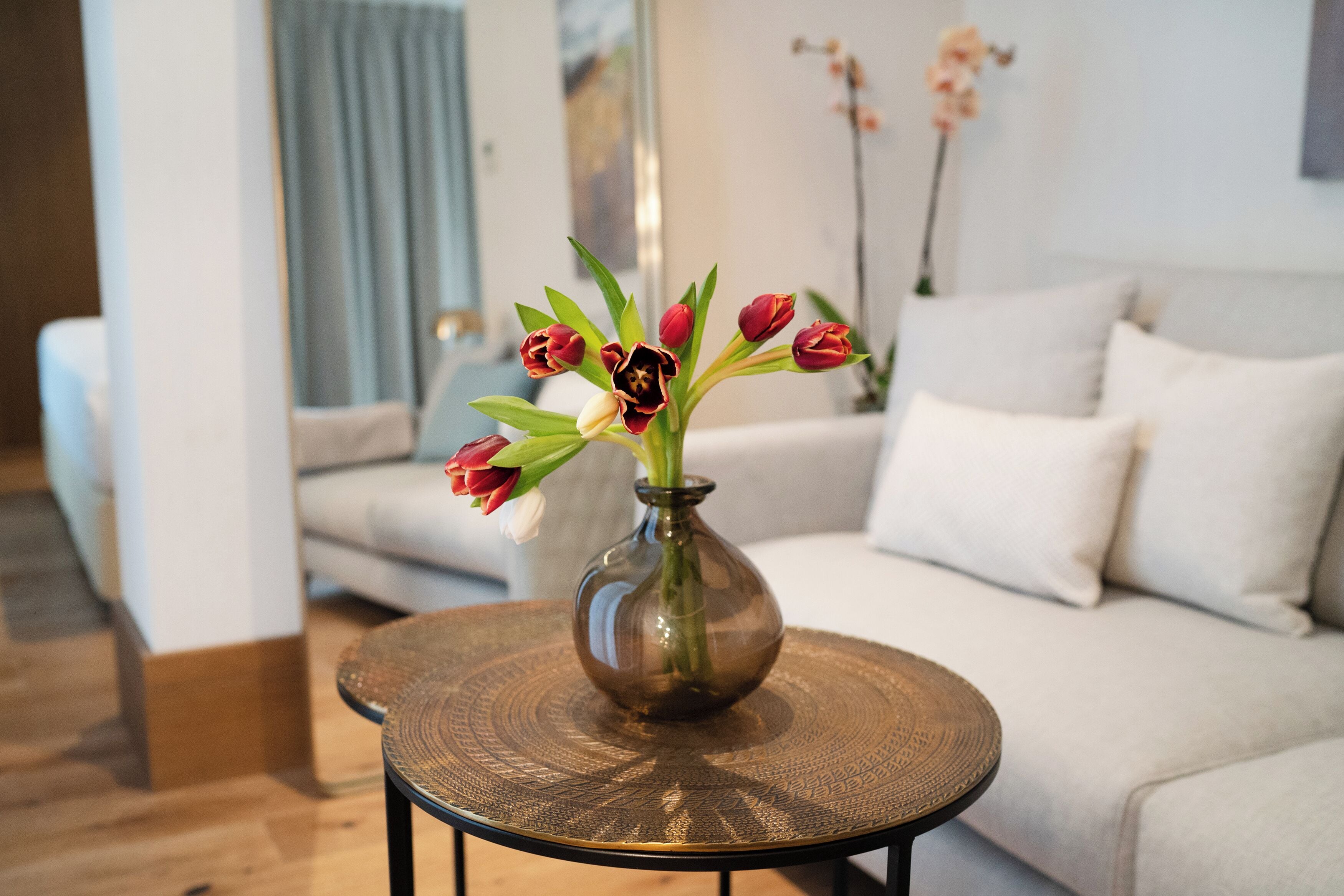 Un salon avec une table ronde en verre, un canapé beige et un vase rempli de fleurs.